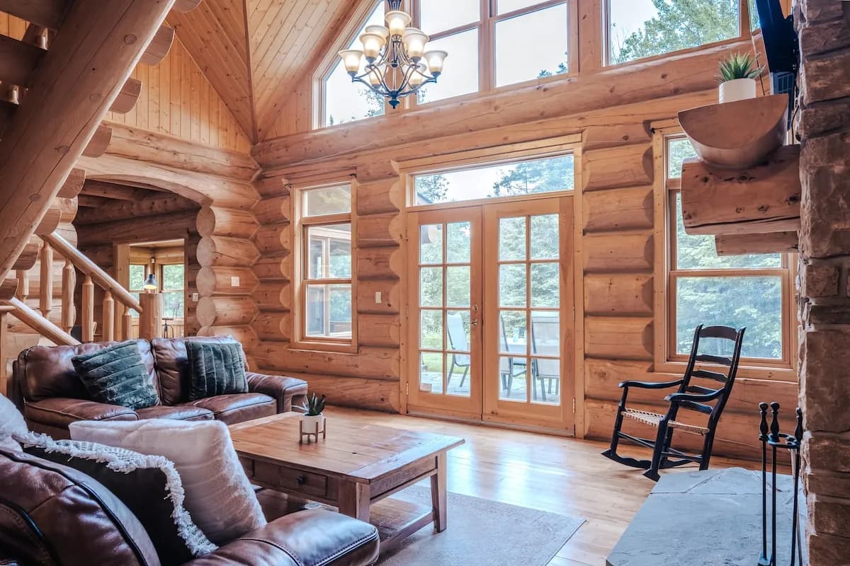 Salon lumineux du chalet en bois rond avec plafond cathédrale et grandes fenêtres orientées plein sud, donnant…